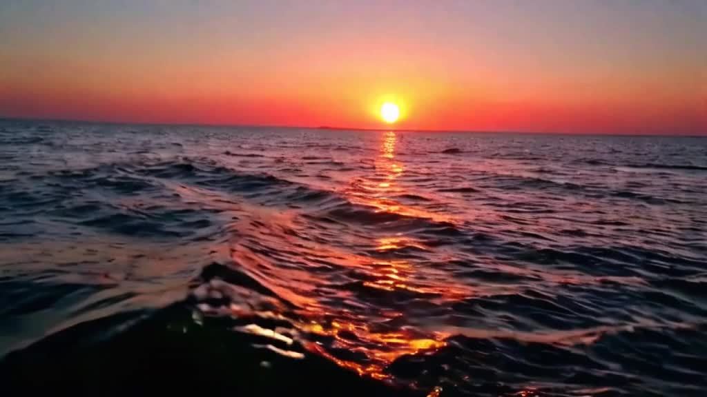 夕阳西下时宁静辽阔海洋的画面。天空被柔和的粉红色和橙色色调所描绘。相机缓缓向下移动至地平线，太阳在那里即将落山。随着光线的消逝，一声最后的“唵”声充满了空间，象征着灵魂永恒的安息。