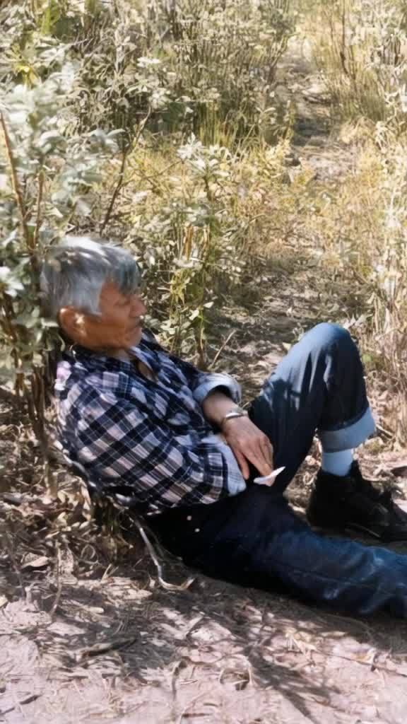 一个人斜靠在灌木丛旁，他穿着格子衬衫和蓝色牛仔裤，