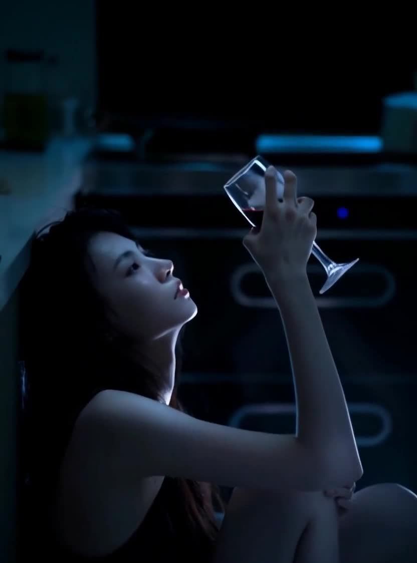 一名女子侧身坐在一个偏暗的房间里，靠着墙。她穿着无