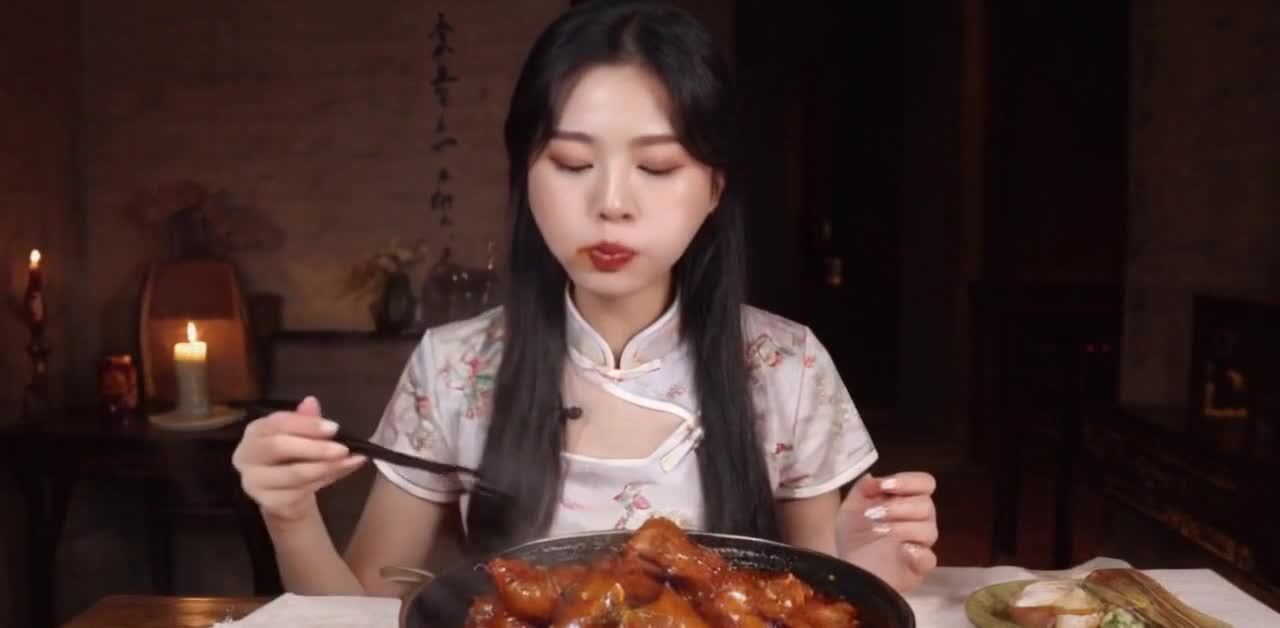一位身穿旗袍的女性坐在桌前，正用筷子夹取一盘红烧肉
